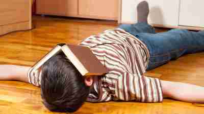 Boy lying on the floor with a book over his face to escape his homework problems
