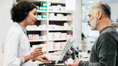 A pharmacist talking to a customer about their prescription, with medications shown in shelves in the background. Many individuals with ADHD have been made to feel like criminals during the stimulant shortage.