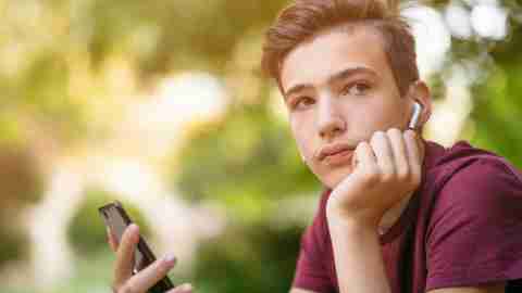 Close-up portrait of a thoughtful unhappy teenage boy with smartphone, outdoors.  Sad teenager with mobile phone looks away, in the park.  Pensive teenager in casual clothes with cell phone in park