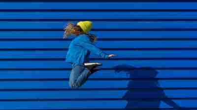Shake loose of your limiting beliefs: A teenage girl wearing a yellow beanie jumping up high against a blue backdrop.
