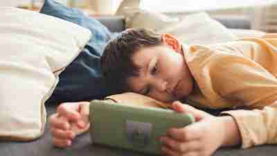 Warm-toned portrait of bored teenage boy using smartphone while lying on bed or couch at home, copy space