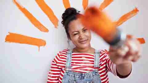 Young woman winking while holding paintbrush against wall during home renovation