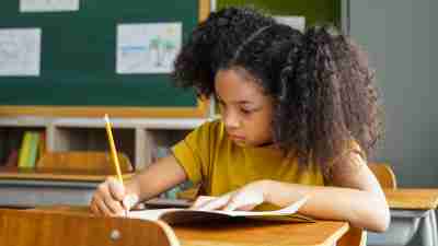 African American school girl sitting in school writing in book with pencil, studying, education, learning. Female student sitting at desk in classroom writing in notebook in exam.