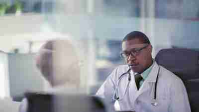 Doctor physician Black man with senior mature adult male man businessman patient sitting with ipad smart tablet computer medical records at desk in sunny bright daylight modern contemporary business hospital office well dressed lab coat planning scheduling care support dedication healthcare medical clinic diagnosis treatment considerations conversation concern asking questions curiosity solutions agreement eye to eye face to face