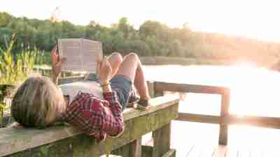 Young girl reading a book while lying on her back on a wooden beam at a wooden dock at the lake.